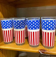 Eight tall glass tumblers with American flag design featuring blue star band and red and white stripes on wooden shelf.