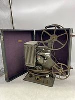 Side angle view of the Keystone 8mm movie projector showing two reels, lens, controls, and coiled power cord. Projector is upright in an open hard carrying case lined with purple fabric.