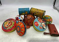 Nine vintage tin noisemakers displayed on a white surface, featuring clown faces, circus performers, and colorful graphics including round, cylindrical, and rectangular shapes.