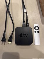 Apple TV box with remote and two cords (power and HDMI) connected and laying on a beige surface.