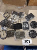 Overview of the belt buckles laid out showing various Guess brand buckles, prong style silver buckle and black decorative buckles, some still wrapped in plastic.