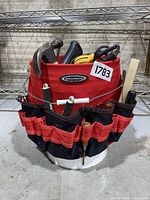 Front side view of red and black fabric tool bucket with multiple small tools visible in outer pockets and some taller tools inside the bucket including hammer and yellow electric sander.
