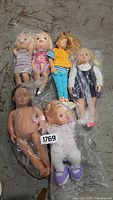 Seven collectible dolls of various sizes and styles, some in plastic wrapping, shown on concrete floor.