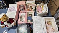 Full lot display showing six collectible dolls in boxes, doll clothes, a teddy bear, and a vintage doll book.
