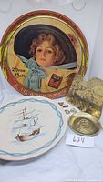 Photo showing a vintage round metal cigarette tin with a young girl illustration and Atlantic Cigarettes branding, ceramic plate with sailing ship design, textured metal elephant figurine, small brass dish, small cufflinks and carved trinkets arranged together.
