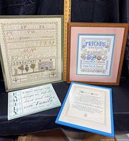 Four framed needlepoint and cross-stitch family themed artworks displayed together against a black backdrop with measuring stick for size reference.