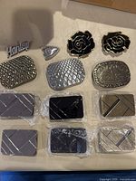 Top view of 13 belt buckles arranged on a surface, showing various textures, shapes, and some still in protective plastic wrapping.