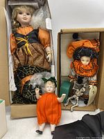 Full view of the three Halloween-themed collector dolls boxed: porcelain doll in orange and black dress, clown doll with ruffled hat, baby doll in pumpkin costume.