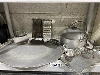 Photo showing entire lot on shelf including large hammered aluminum tray with decorative edge, two flat choppers, two metal graters and aluminum cooking pot with lid and handle