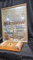 Framed Hennessy cognac mirror with gold decorative grapevines and Hennessy logo, photographed against a black fabric background, with Buddha clock in front.