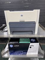 Front view of the beige and gray HP LaserJet 1320n printer on a metal table. The HP 49X Laserjet toner cartridge in brand box is placed underneath.