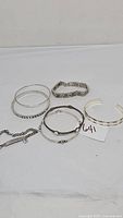 Group of five silver tone bracelets displayed on a table, showing various styles including bangles, chain, and cuff with floral designs.