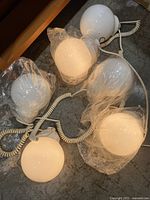 Six white plastic hanging globe lights with coiled white electrical cords, some globes wrapped in plastic, arranged on floor surface.