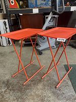 Two mid century style orange plastic top dinner trays with matching orange metal foldable legs, shown on a concrete floor in an indoor setting.