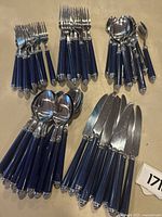 Full view of approx 50-60 pieces of Mikasa flatware split into forks, knives, spoons, and teaspoons with blue handles and stainless steel heads arranged on flat surface.