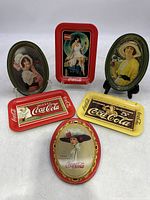 Six miniature Coca-Cola advertising tin trays of various shapes and vintage artwork featuring women with Coca-Cola branding.
