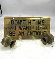 Brass novelty license plate with black lettering reading 'DON'T HIT ME I WANT TO BE AN ANTIQUE' displayed with two antique horn noisemakers, showcasing wear and age.