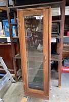 Front view of tall oak wood display cabinet with glass door closed, showing wood frame and clear glass panel.