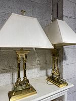 Pair of brass metal table lamps with rectangular cream shades placed side by side against a concrete block wall