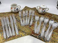 Complete lot showing 4 fabric placemats underneath sets of stainless steel cutlery (knives, forks, spoons) all wrapped in plastic, and 2 handpainted ceramic containers (cream and sugar) with rooster/chicken designs.