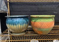 Two ceramic planter pots placed side by side on a metal shelf outdoors. The left pot is glazed in blue and cream gradient, the right pot is terra-cotta with green and orange decorative patterns and visible chipping.
