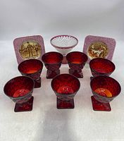 Front view of seven vintage deep red glass goblets with diamond cut pattern and square bases, two character saucers behind, and a diamond cut clear vase with cranberry rim behind