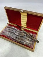 Wooden box open showing six steak knives wrapped in plastic with red felt interior.