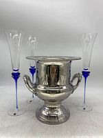 Overall view of the Leonard silver plated ice bucket with three blue stemmed champagne flutes around it on a white surface.