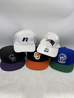 Front view of five sports hats showing New England Patriots white hat, white hat with blue R, Colorado Rockies, CU, and blue/green wolf logo hats