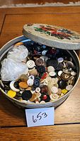 Open vintage tin showing variety of buttons inside, including white, black, clear, yellow, brown, and some patterned buttons.