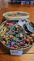 Open vintage metal tin containing colorful assorted costume jewelry including beaded necklaces and metal bangles.