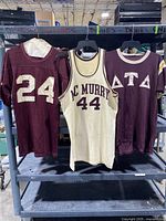 Three vintage shirts hanging on a rack: a burgundy jersey with number 24 on left, a cream basketball jersey with MC Murry 44 in center, and a burgundy Delta fraternity shirt on right.