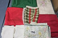 Photo showing folded linens including solid green, red, white napkins, floral print, checkered cloth with lace trim, red textured cloth, and off-white embroidered linen.