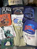 Top view of 10 vintage destination graphic t-shirts folded and stacked showing various location graphics.