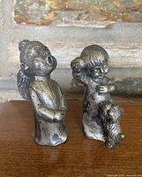 Front view of two pewter figurines on wooden surface with brick background; singing angel on left, girl with dog on right.