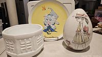Photo showing three porcelain decorative items, a white planter, egg-shaped figurine, and a square plate with stylized girl illustration