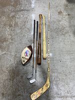 Lot of vintage sports equipment on concrete floor showing wooden hockey stick with multiple autographs, two wooden baseball bats, two golf clubs, and a Buffalo Bills football.