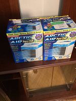 Two unopened boxed Arctic Air Pure Chill evaporative air coolers side by side on a wooden surface.