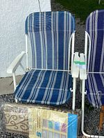 Two blue striped foldable lawn chairs with white metal frames, outdoor rug packaged in plastic leaning against the chair.
