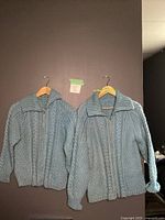 Two light blue hand-knit cable pattern wool sweaters hanging side by side, full front zippers, ribbed cuffs and collars visible.