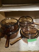 Five amber glass cookware pieces with handles and matching lids arranged inside a cabinet shelf.