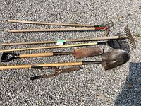 Photo displaying seven garden and outdoor tools laid on gravel surface, showing full length and condition of wooden handles and metal heads