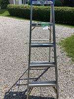 Front view of aluminum step ladder standing on gravel outdoors, showing five steps and blue top.
