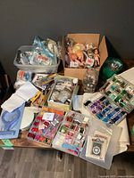 Overview of craft supplies spread on table including embroidery threads, boxes of dolls, and jars of buttons