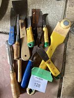 Photo showing assortment of scrapers, putty tool, and cutting utensils with wood and plastic handles in various colors. Visible rust and wear on blades and handles.