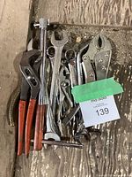 Full view of assorted metal hand tools laid out on wooden floor, including various wrenches, pliers with red handles, locking pliers with curved jaws, needle-nose pliers, and a socket wrench with wooden handle.