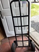 Front view of black metal hand truck leaning against a white door, showing straight handle and metal frame.