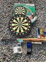 Photo showing dartboard with box, wood Cooper baseball bat, and three sets of darts with accessories laid out on gravel
