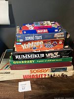 Stack of 9 boxed family board games piled on top of each other against a dark wall with visible wear on boxes