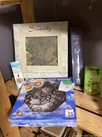 Photo showing the Serenity stepping stone wall plaque, Crystal Twister box, Wind Spinner box, and Garden Butterfly lights box on a wooden shelf.